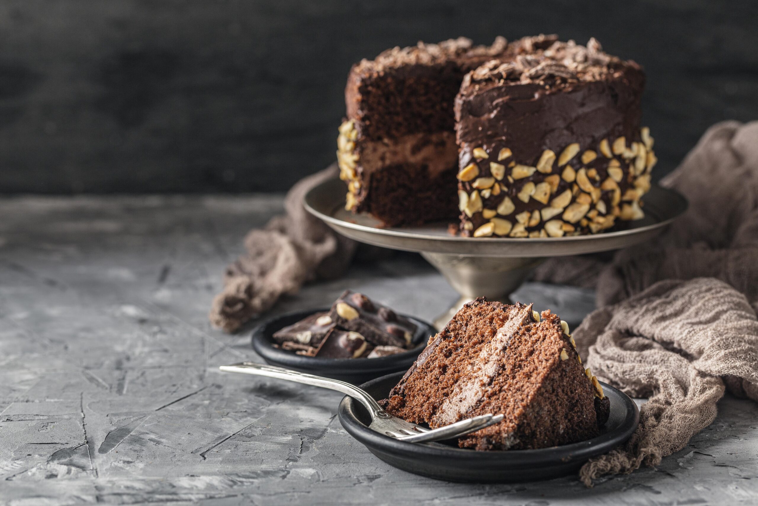 A beautifully styled German Chocolate Cake displayed on a white ceramic cake stand. The cake features three rich chocolate layers with visible coconut-pecan frosting between each layer and on top. Decorative pecans and shredded coconut are sprinkled over the frosting, creating a rustic yet elegant look. The scene is set in a softly blurred kitchen background with warm, natural light adding a cozy atmosphere